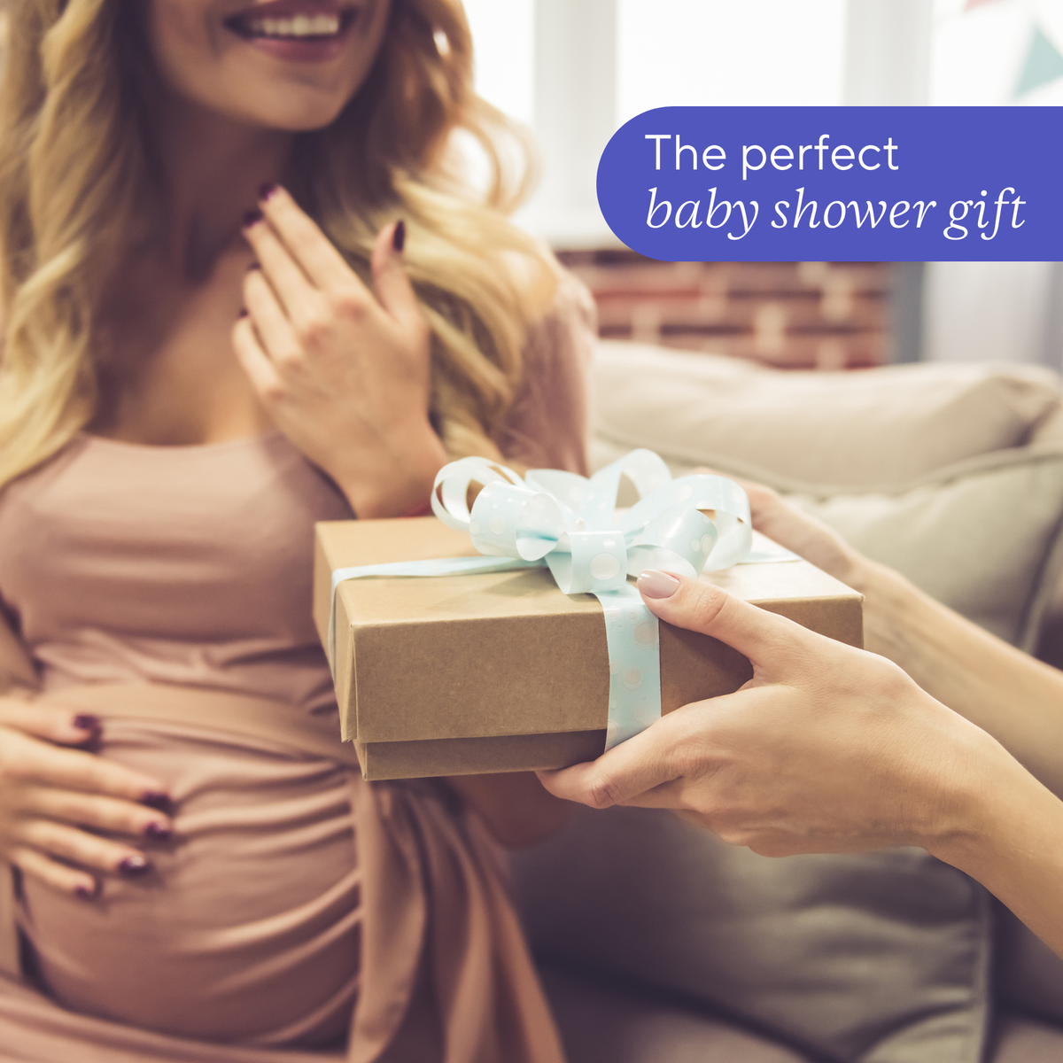 A person in a dusty pink maternity gown receives a brown kraft gift box tied with a pale blue ribbon, with a purple text badge reading 'The perfect baby shower gift' in the upper right.